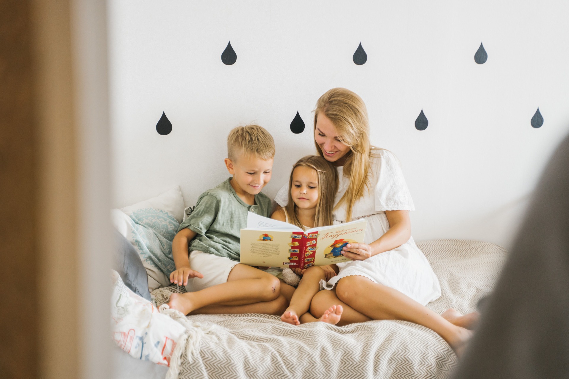 Äiti ja kaksi lasta lukevat kirjaa sängyllä, taustalla mustia vesipisara-kuvioita.